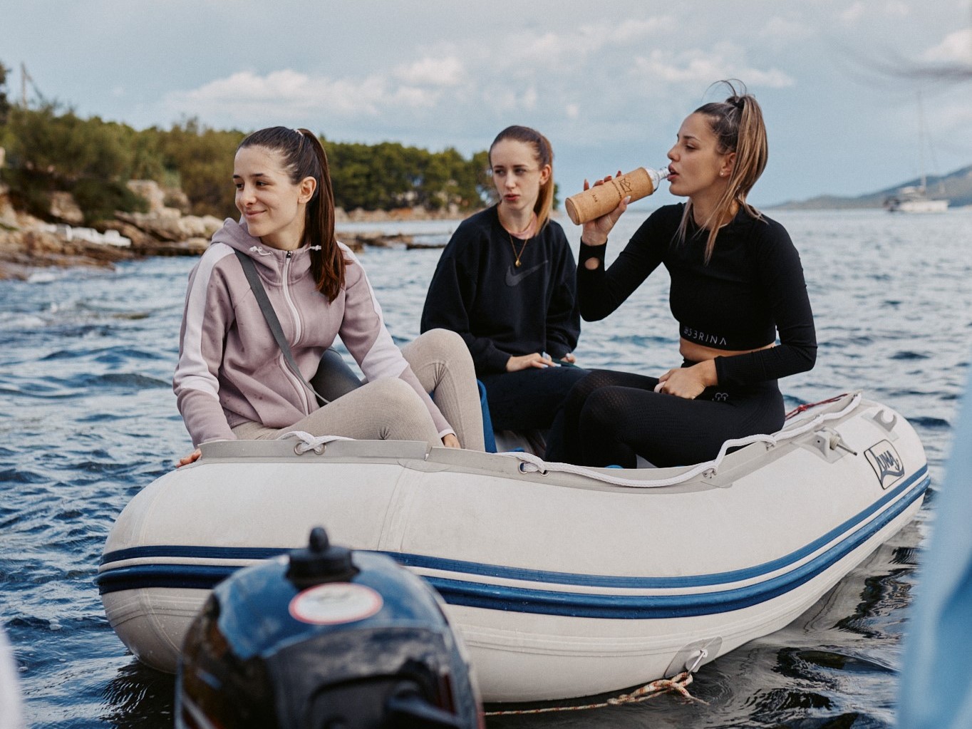 Three women in sportswear sit on an inflatable boat; one drinks from a cork-sleeved glass bottle; calm sea and rocky shore in the background.